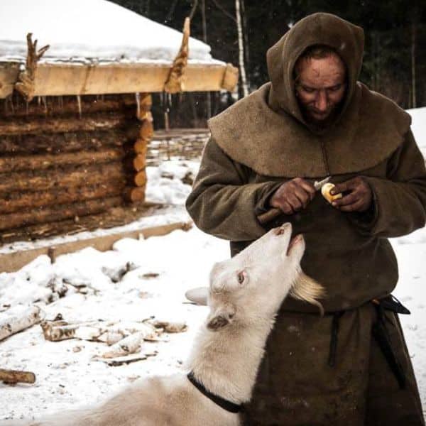 A peasant in a rough brown cloak stands in the snow peeling a root vegetable, while a white goat eagerly looks up at him, with a rustic log shelter in the background.