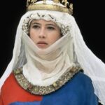 A cinematic portrayal of Isabella of France from the film Braveheart, featuring a woman in regal medieval attire. She wears a white veil, a golden crown, and an ornate embroidered necklace, representing her noble lineage and powerful historical presence.