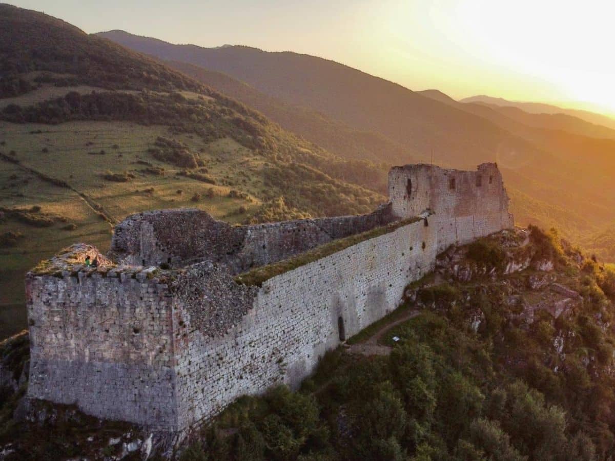 An aerial view of Montségur fortress at sunset, its crumbling walls glowing warmly as rolling hills and valleys stretch into the horizon.