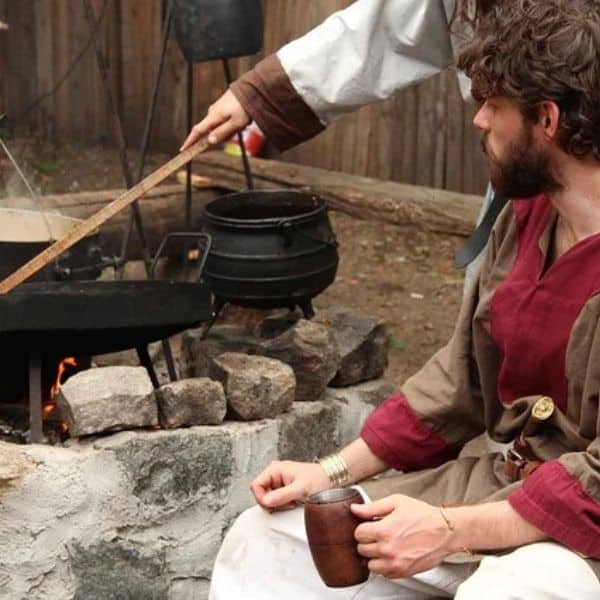 A man in a simple red and brown tunic holds a wooden mug near a fire pit, watching as food cooks in cast iron pots hanging over the flames.