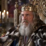 A regal man with long gray hair and a full beard sits on an ornate throne, wearing a golden crown and a fur-trimmed robe, symbolizing the forgotten grandeur of medieval monarchs.