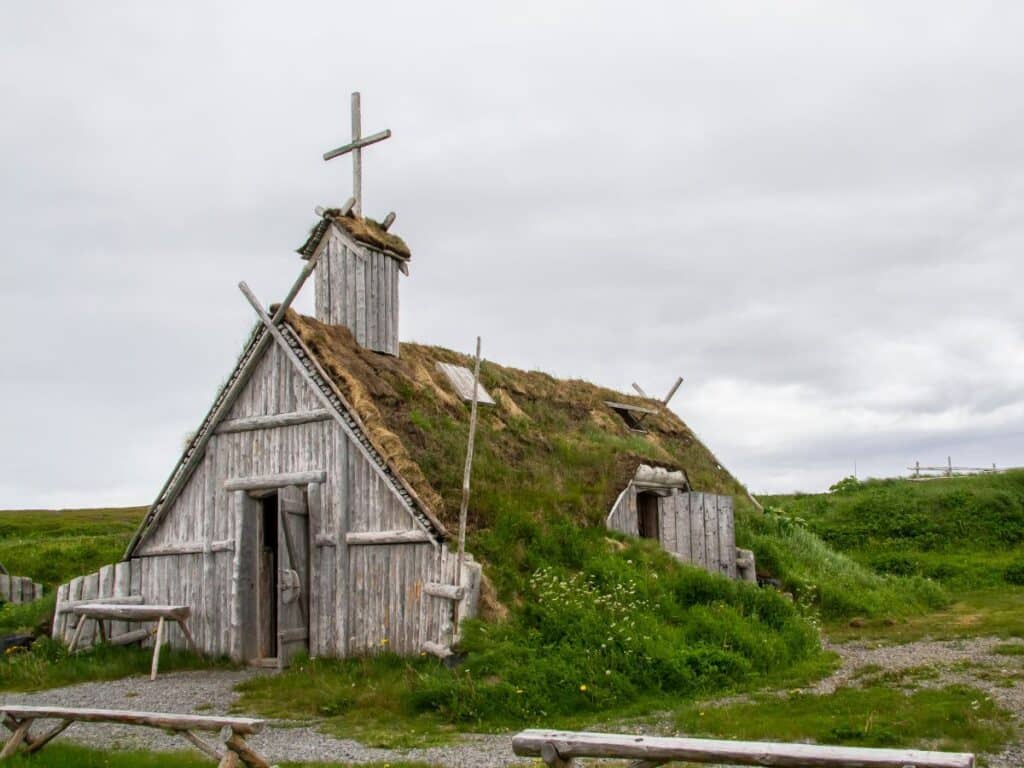 A reconstructed Viking sod house with a wooden frame and cross atop the roof, surrounded by lush green grass under a cloudy sky, depicting early Norse settlement life.