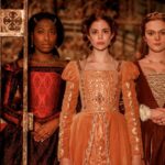 Dramatic still from "The Spanish Princess" featuring Catherine of Aragon, Bessie Blount, and another lady-in-waiting dressed in regal Tudor gowns, standing behind palace gates.