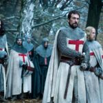 Live-action reenactment of Templar knights in a forest from the Knightfall series, showcasing chainmail armor, red crosses on white capes, and a solemn tone reflective of the legacy of the Knights Templars in pop culture.