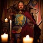 A modern cinematic portrayal of King Arthur sitting sternly on a throne in candlelight, dressed in a lion-emblazoned tunic and red cape, gripping a sword with both hands.