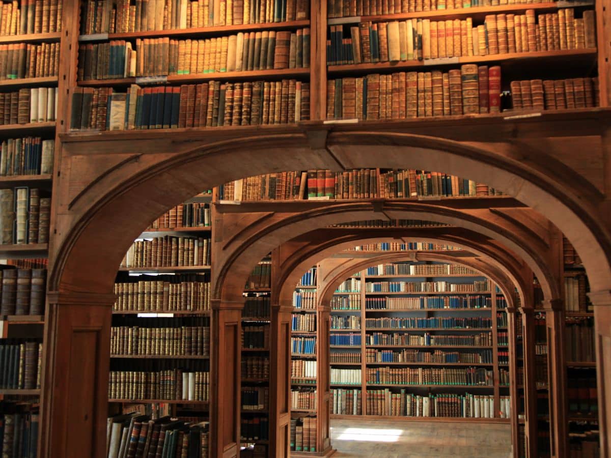 View of an ornate historical library filled with tall wooden bookshelves and arched wooden supports, evoking the scholarly depth behind many books on the Hundred Years War and medieval studies.