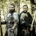 Two reenactors dressed as medieval warriors in the Hundred Years War, standing in a forest with intense expressions; one holds a battle axe and wears chainmail and scale armor, while the other grips a longsword and wears studded leather and metal shoulder guards.
