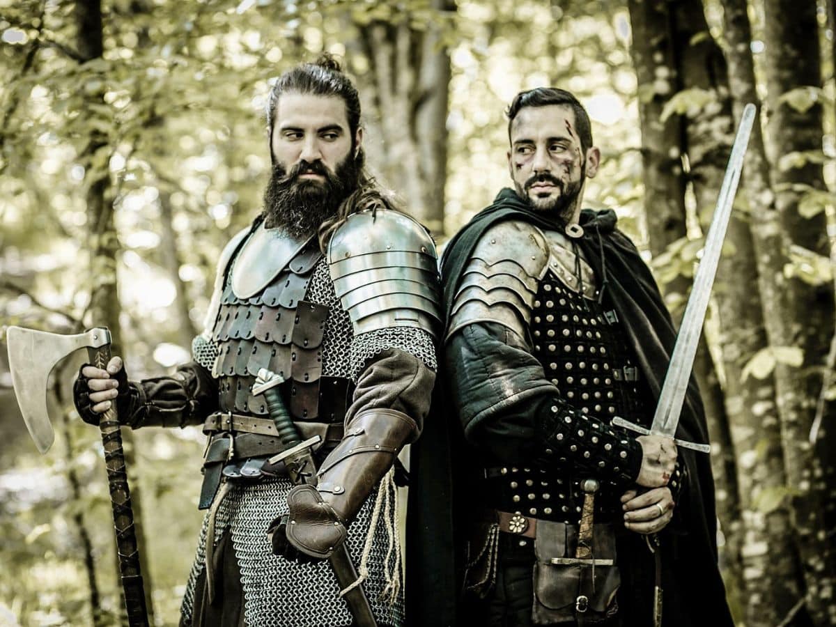 Two reenactors dressed as medieval warriors in the Hundred Years War, standing in a forest with intense expressions; one holds a battle axe and wears chainmail and scale armor, while the other grips a longsword and wears studded leather and metal shoulder guards.