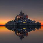 Silhouette of Mont Saint-Michel at dusk reflected in still water, its medieval spires and glowing lights contrasting against a fiery orange and purple sky.
