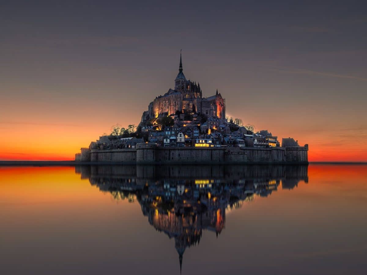 Silhouette of Mont Saint-Michel at dusk reflected in still water, its medieval spires and glowing lights contrasting against a fiery orange and purple sky.