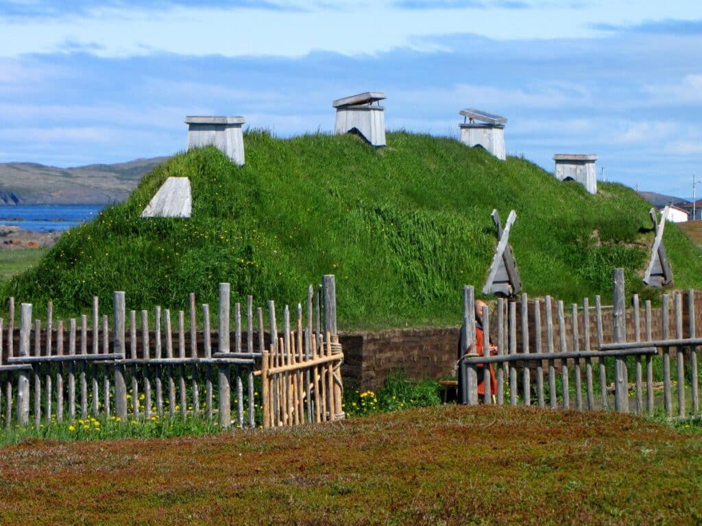 A reconstructed Viking settlement with sod-covered longhouses and wooden fences, set by a coastline, representing archaeological evidence in the debate over "Did the Vikings Really Sail to America."