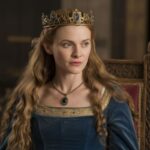 Actress portraying Empress Matilda seated on a throne, wearing a blue velvet gown with a golden crown and necklace, symbolizing her claim to the English throne and royal status.