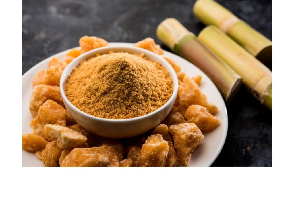 A white plate filled with jaggery chunks and a bowl of powdered jaggery sits beside sugarcane—illustrating how sweeteners like cane sugar and jaggery were treasured ingredients in Middle Ages desserts.