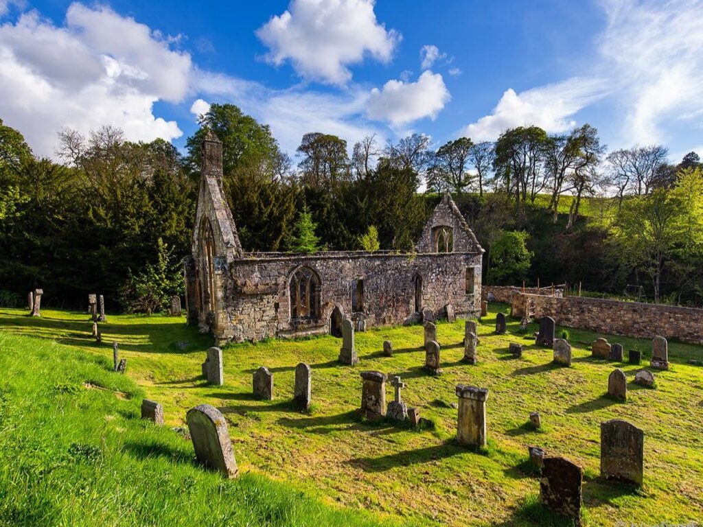 Scenic view of a ruined medieval chapel surrounded by gravestones in a grassy graveyard, possibly linked to the Knights Templar in England, nestled in the English countryside.