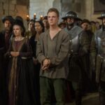 A scene from a historical TV series depicting Perkin Warbeck in plain clothing, surrounded by armored guards and noble spectators, during his public downfall and trial under Henry VII.