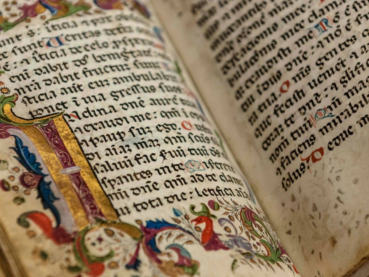 Close-up of a medieval illuminated manuscript with ornate gold and colored illustrations, showing Latin text in black ink with decorative initials and red rubrication, symbolizing the era when many now-common medieval sayings originated in religious and literary works.