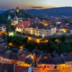 Aerial view of Sighisoara at dusk, a well-preserved medieval European citadel in France, showcasing fortified walls, towers, and glowing streets nestled in the hills.