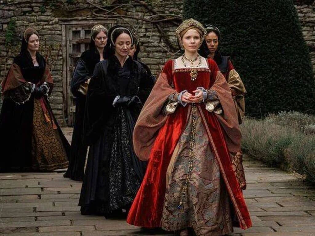 A dramatic scene from Wolf Hall shows Jane Seymour dressed in rich red and gold Tudor attire, walking solemnly with her ladies-in-waiting in a stone courtyard, capturing the elegance and restraint associated with Jane Seymour's character.