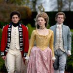 Promotional still from a period drama showing three aristocratic characters walking confidently through formal gardens—Lady Worsley in a golden gown and two men in 18th-century attire. The image evokes the intrigue and scandal tied to Lady Worsley's real-life story.