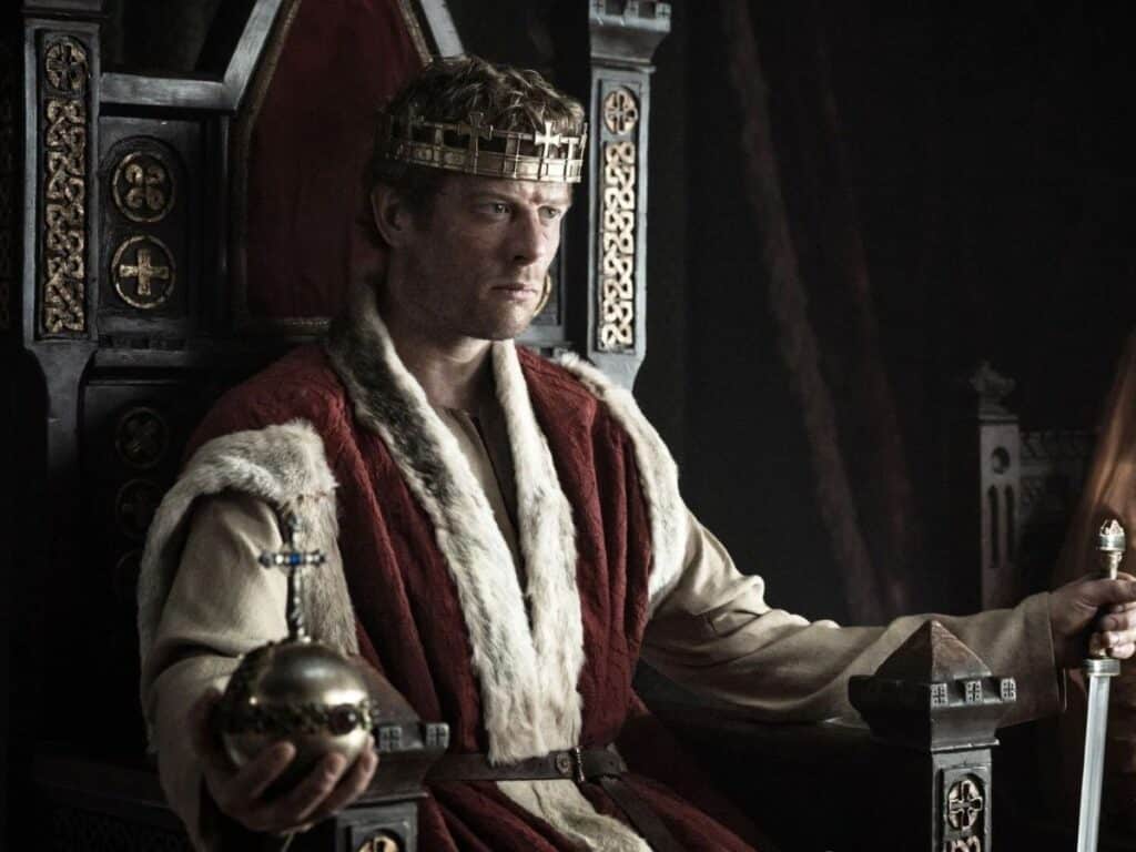 A solemn king in medieval robes and crown sits on a gothic throne holding a scepter and orb, capturing the royal atmosphere of a Middle Ages TV series.