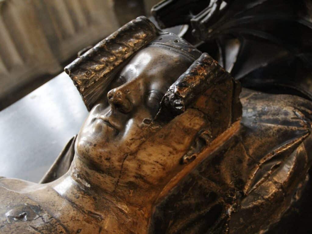 Close-up of the tomb effigy of Philippa of Hainault, showing a weathered but serene face framed by a crown and elaborate headpiece. The bronze sculpture lies in Westminster Abbey, commemorating the queen consort’s legacy.