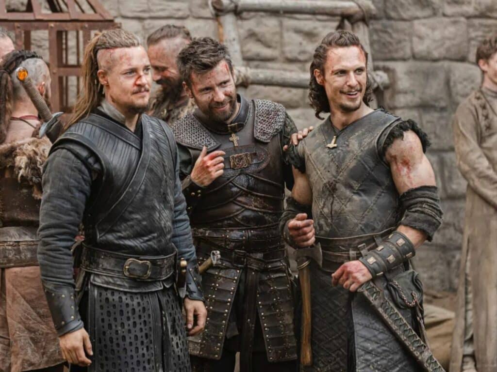Three battered warriors in leather and chainmail smile post-battle in a medieval courtyard, showing camaraderie in a gritty Middle Ages action series.