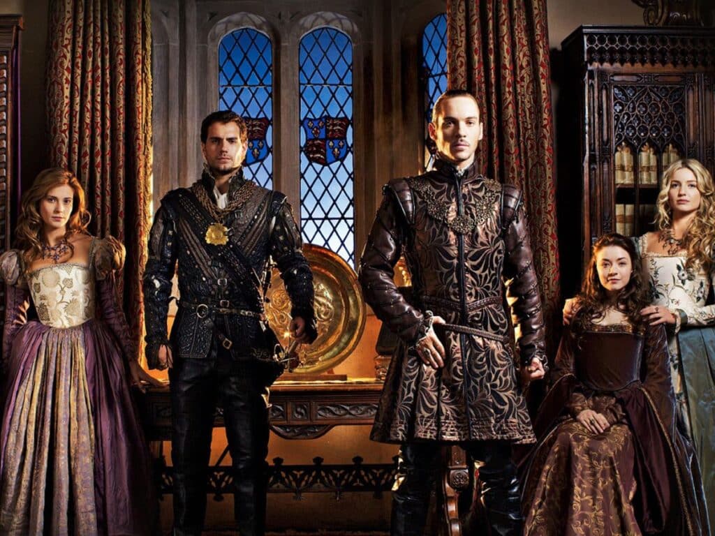 A noble group in elaborate Renaissance outfits poses in a lavish chamber with stained-glass windows, portraying court life in a TV series about the Middle Ages.