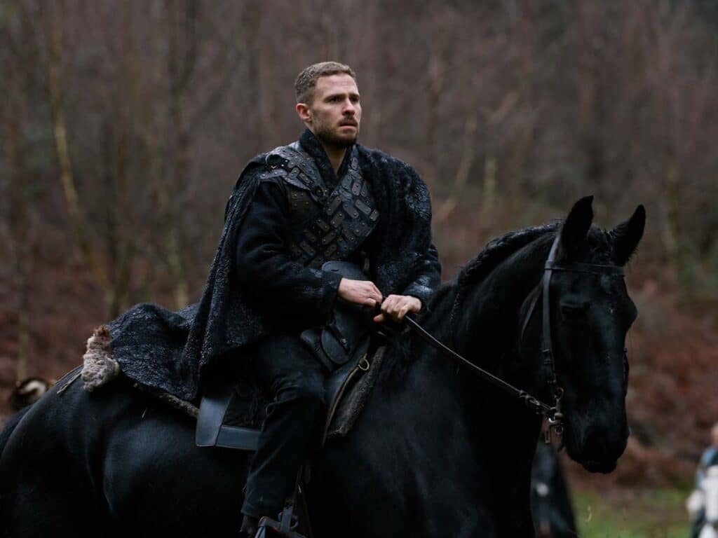 A brooding warrior in black medieval armor rides a dark horse through a bleak, wintry forest, capturing the gritty atmosphere of TV shows set in the Middle Ages.