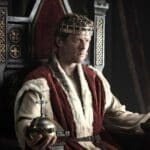 A medieval king wearing a golden crown and fur-lined robe sits on an ornate throne, holding a royal orb in his hand. The scene captures a solemn moment of power in King and Conqueror.