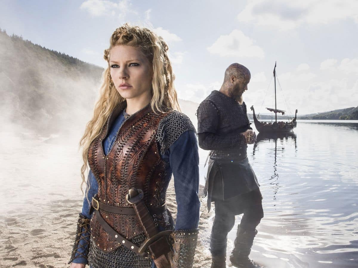 A blonde woman dressed in detailed leather armor stands on a misty shoreline, looking determined, while a man in black armor stands behind her near a Viking ship on calm waters. This scene portrays Lagertha in a dramatic Norse setting.