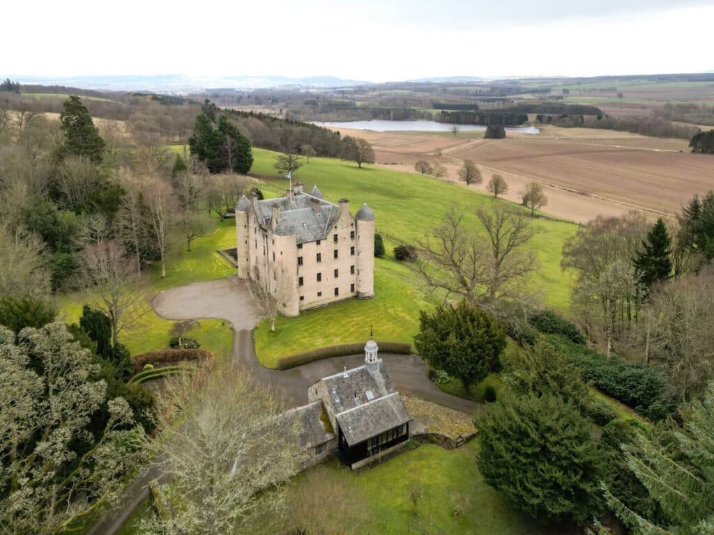 Aerial view of a Scottish countryside estate featuring a turreted stone castle surrounded by farmland and forest, similar to locations associated with Margaret Tudor’s life in Scotland.