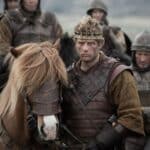 A gritty scene from a historical reenactment or film depicting Harold Godwinson in battle-worn armor, holding the reins of a horse wearing decorative leather headgear, with fellow soldiers standing in the background.