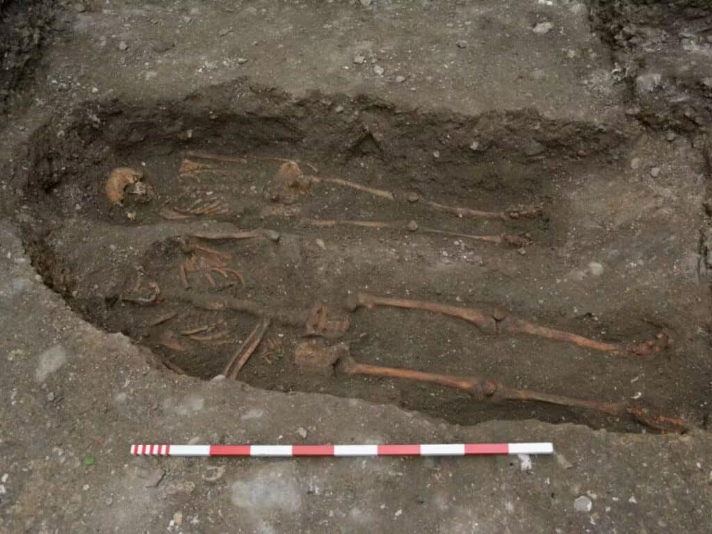 Two human skeletons partially uncovered in an archaeological dig, laid side by side in a single grave, with a measuring rod at the bottom for scale. The burial is believed to be connected to historical findings near the Tower of London.