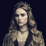 Dramatic portrait of a modern actress portraying Cecily of York, wearing a gold-trimmed gown and delicate crown with pearl-like orbs. Her loose, wavy blonde hair and composed expression evoke royal elegance against a dark background.