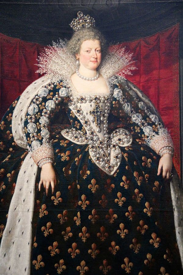 Marie de’ Medici in a grand ceremonial gown adorned with pearls and fleur-de-lis patterns, wearing a crown and standing before a red backdrop