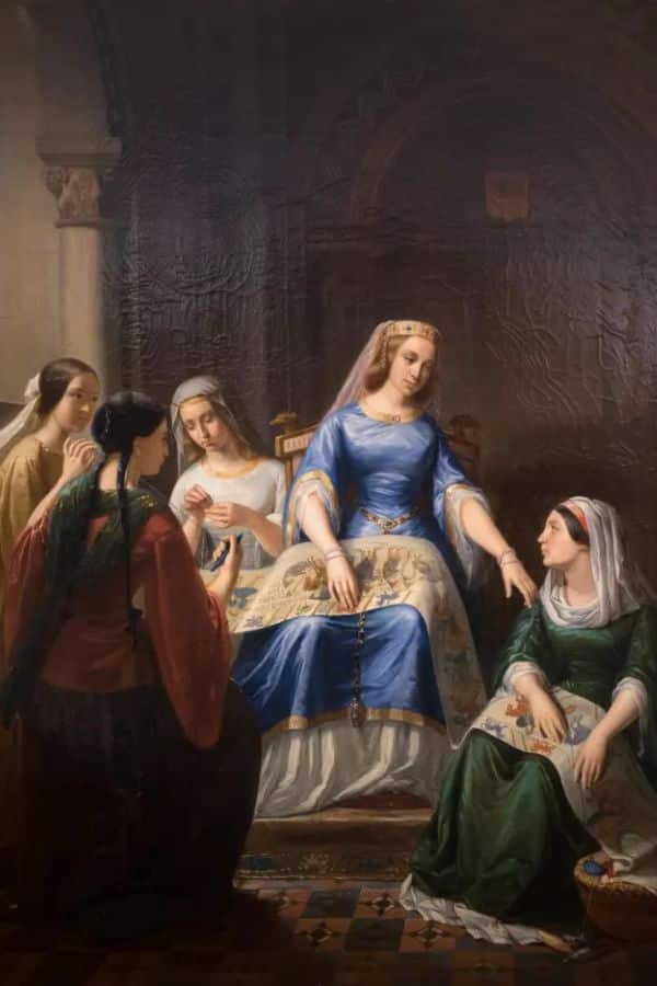 A historical painting depicting Matilda of Flanders seated in a blue and gold gown, surrounded by four women engaged in embroidery, likely referencing the creation of the Bayeux Tapestry. The scene emphasizes Matilda’s royal status and patronage of the arts.