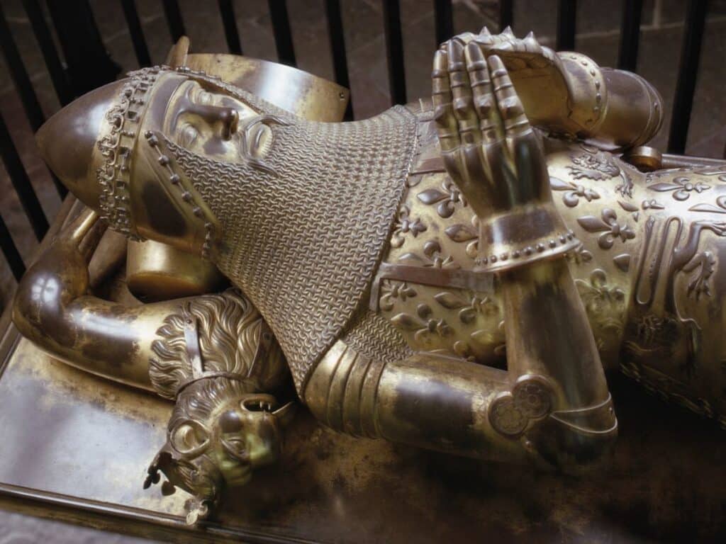 Tomb effigy of the Black Prince in Canterbury Cathedral, showing detailed golden armor with fleur-de-lis patterns and a chainmail coif, hands clasped in prayer.