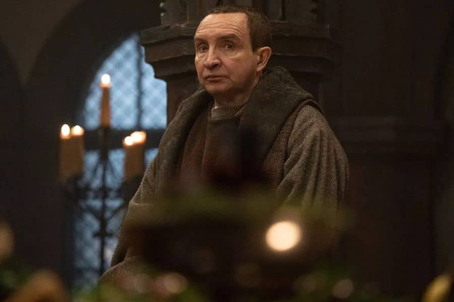 A solemn monk-like figure sits in a candlelit stone chamber, draped in a brown cloak. Character study from BBC One’s new historical drama, King & Conqueror.
