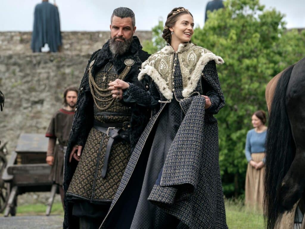 A noble couple in medieval costume walk hand in hand outside a stone wall, the woman smiling in a patterned cloak with fur trim, possibly portraying Emma of Normandy.