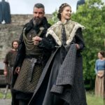 A noble couple in medieval costume walk hand in hand outside a stone wall, the woman smiling in a patterned cloak with fur trim, possibly portraying Emma of Normandy.