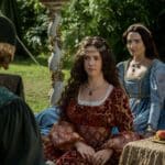 Dramatic reenactment scene from a historical drama featuring an actress portraying Lady Catherine Gordon seated outdoors in an ornate red and gold gown with a jeweled headpiece, flanked by women in Tudor-style dresses with a fruit-laden table nearby.