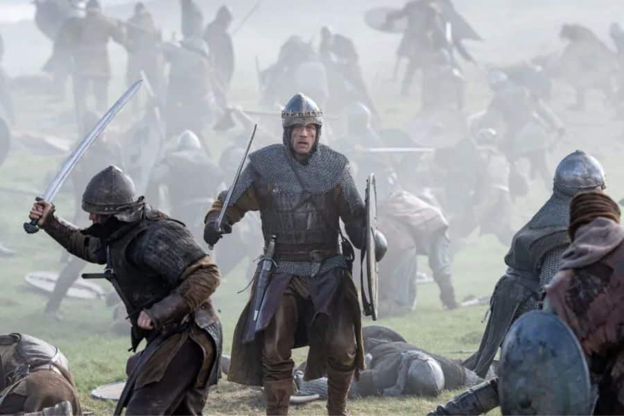 Armored soldiers clash in a foggy battlefield as one central warrior charges forward with sword and shield raised. Action sequence from BBC One’s new historical drama, King & Conqueror.