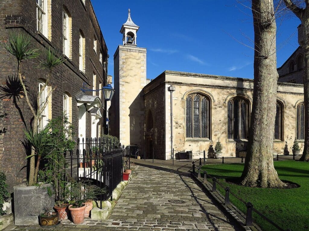 Sunlit exterior view of the Chapel Royal of St Peter ad Vincula, located within the Tower of London, showing its stone walls, arched windows, and adjacent buildings lining a cobbled path. This chapel is the final resting place of many executed Tudor-era figures.