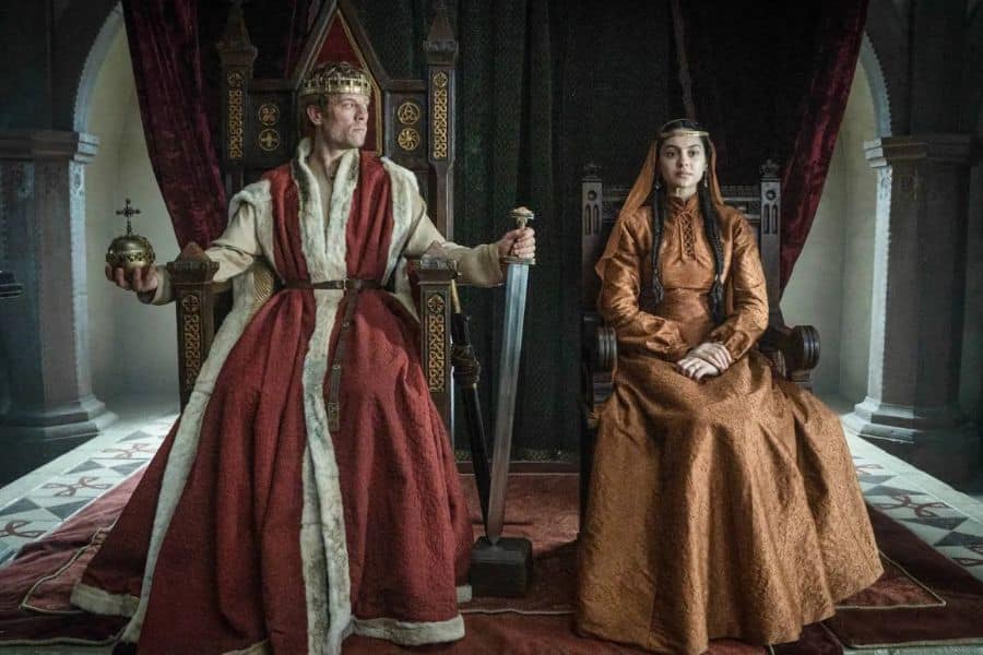 A regal man in a fur-lined red robe and crown sits on a throne holding a sword and orb beside a solemnly seated woman in gold. Royal court moment from BBC One’s new historical drama, King & Conqueror.
