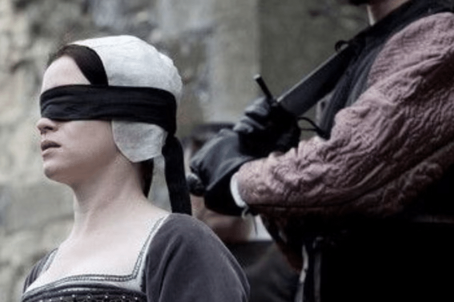 A woman in Tudor-style clothing stands blindfolded, preparing for execution as a man in period attire holds a sword behind her. This dramatized depiction highlights the somber and ritualized nature of Tudor executions.