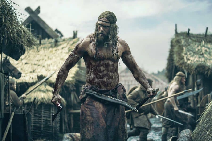 A muscular, blood-covered Viking warrior stands shirtless in a muddy village, gripping a hatchet and sword, with long hair and a fierce expression. Behind him, other warriors move through the thatched-roof settlement, suggesting the aftermath of a brutal battle.