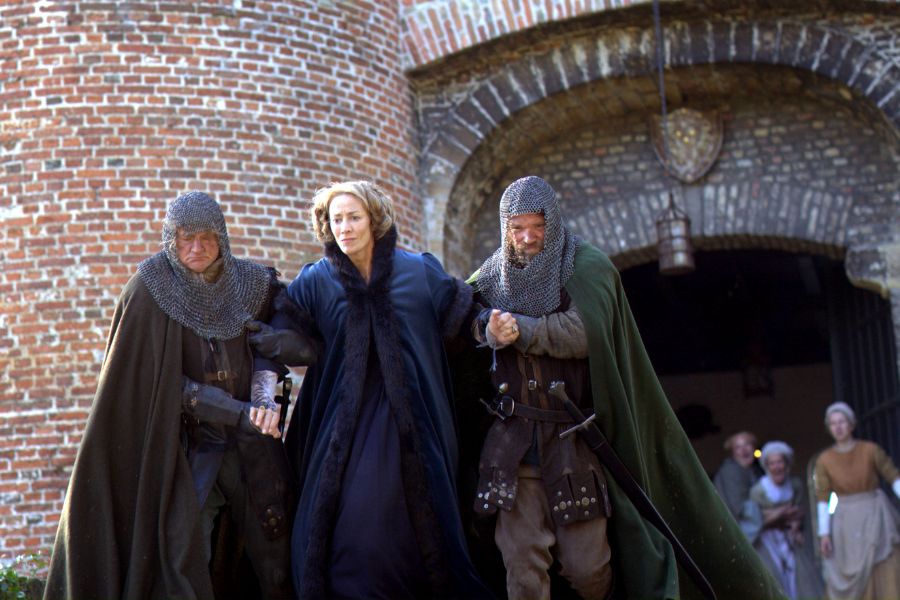 Jacquetta of Luxembourg, in a fur-lined blue cloak, is escorted by two armored guards outside a medieval castle gate, with onlookers watching from the background.