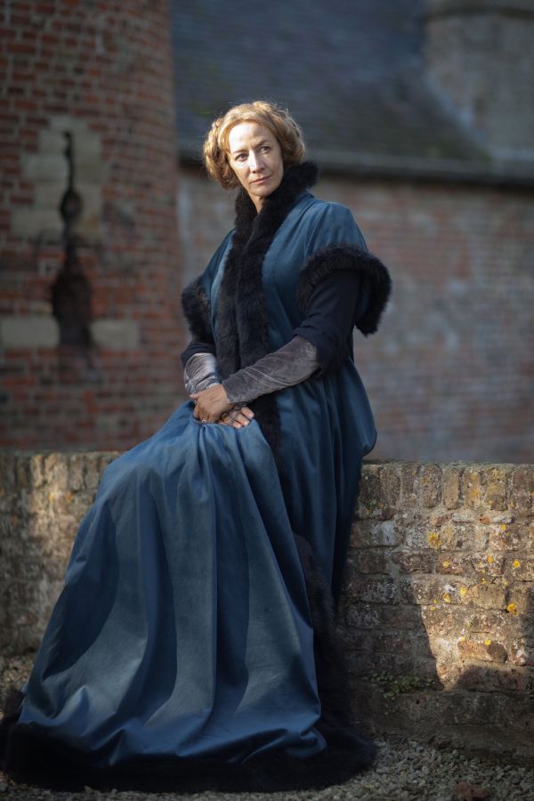 A woman dressed as Jacquetta of Luxembourg in a deep blue gown trimmed with black fur sits gracefully on a stone wall, with a medieval brick structure in the background.