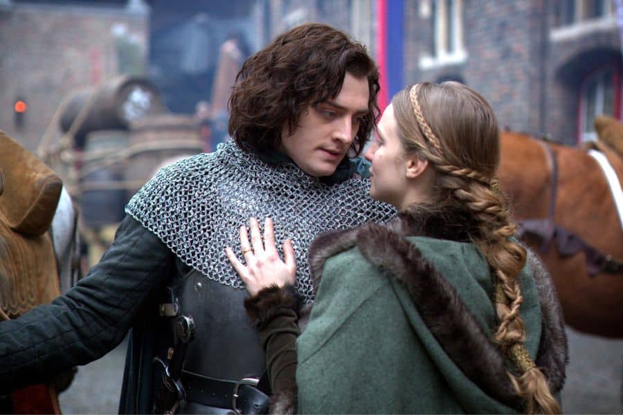 Scene from a historical drama portraying Anne Neville and Richard III in a tender moment, dressed in medieval-style clothing with a castle setting behind them.