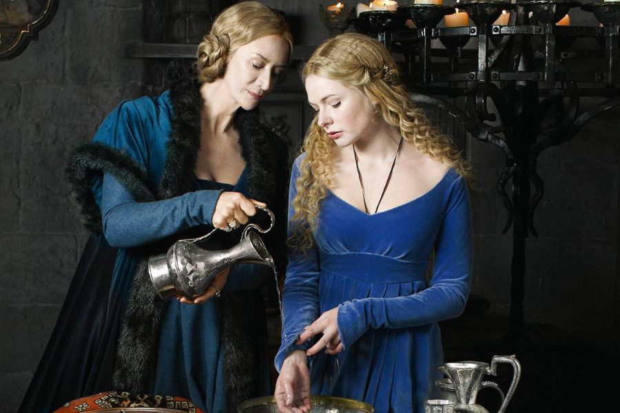 Jacquetta of Luxembourg, wearing a teal gown, gently pours water from a silver pitcher as a young blonde woman in a blue dress washes her hands in a candlelit medieval setting.